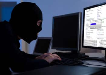 Masked hacker wearing a balaclava sitting at a desk downloading private information off a computer. Avoiding fraud.