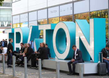 Attendees sit beside a Traton logo outside the Volkswagen AG (VW) exhibition area at the IAA Commercial Vehicles Show in Hanover, Germany, on Wednesday, Sept. 19, 2018. Volkswagen AG's truck unit is pushing to reduce its dependence on its main European market and lift profit margins in a challenge to commercial-vehicle industry leaders Daimler AG and Volvo AB.